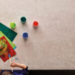 Caesarstone 4023 Topus Concrete - Children paint using colorful paints and brushes, creating art on paper. Three paint containers are nearby on a light-colored surface in a creative setting.
