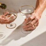 Caesarstone 5140 Dreamy Carrara - Champagne bottle pouring into two coupe glasses, held by two hands on a sunlit marble surface, with dried flowers nearby.
